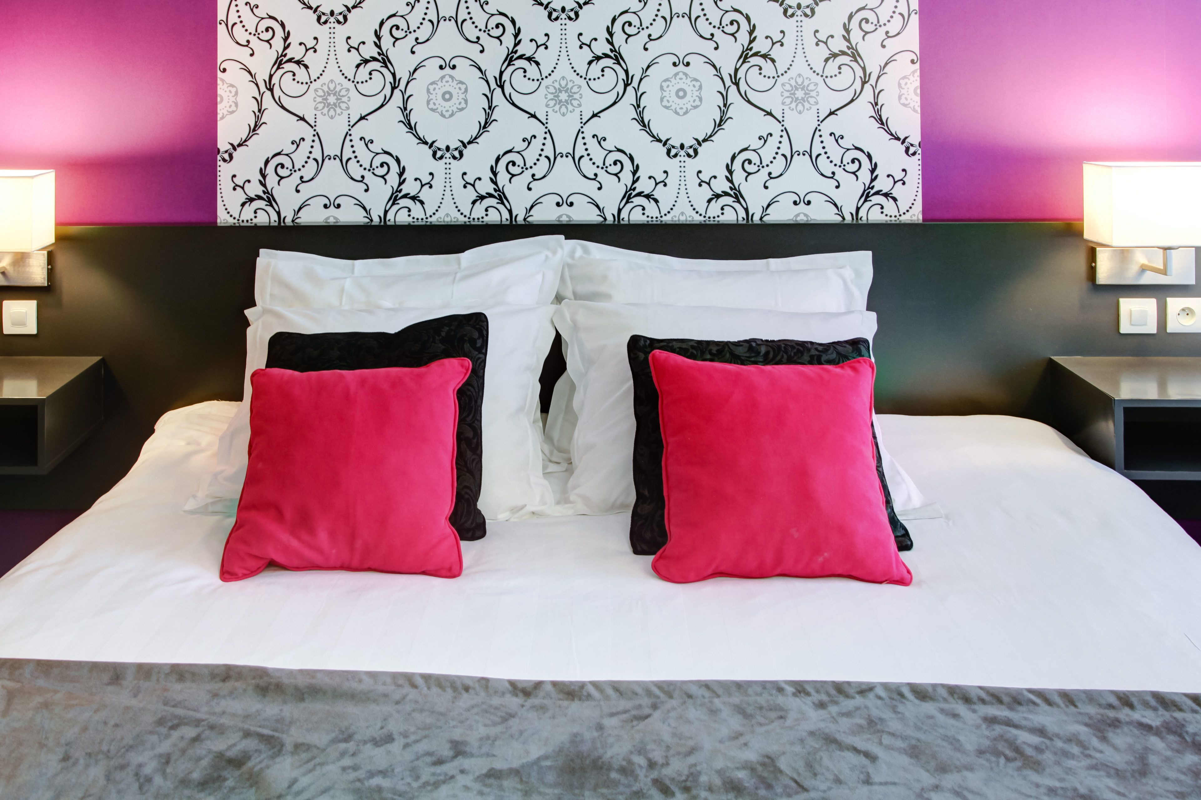 A bed with white and black bedding, two red pillows, and a patterned wallpaper above.