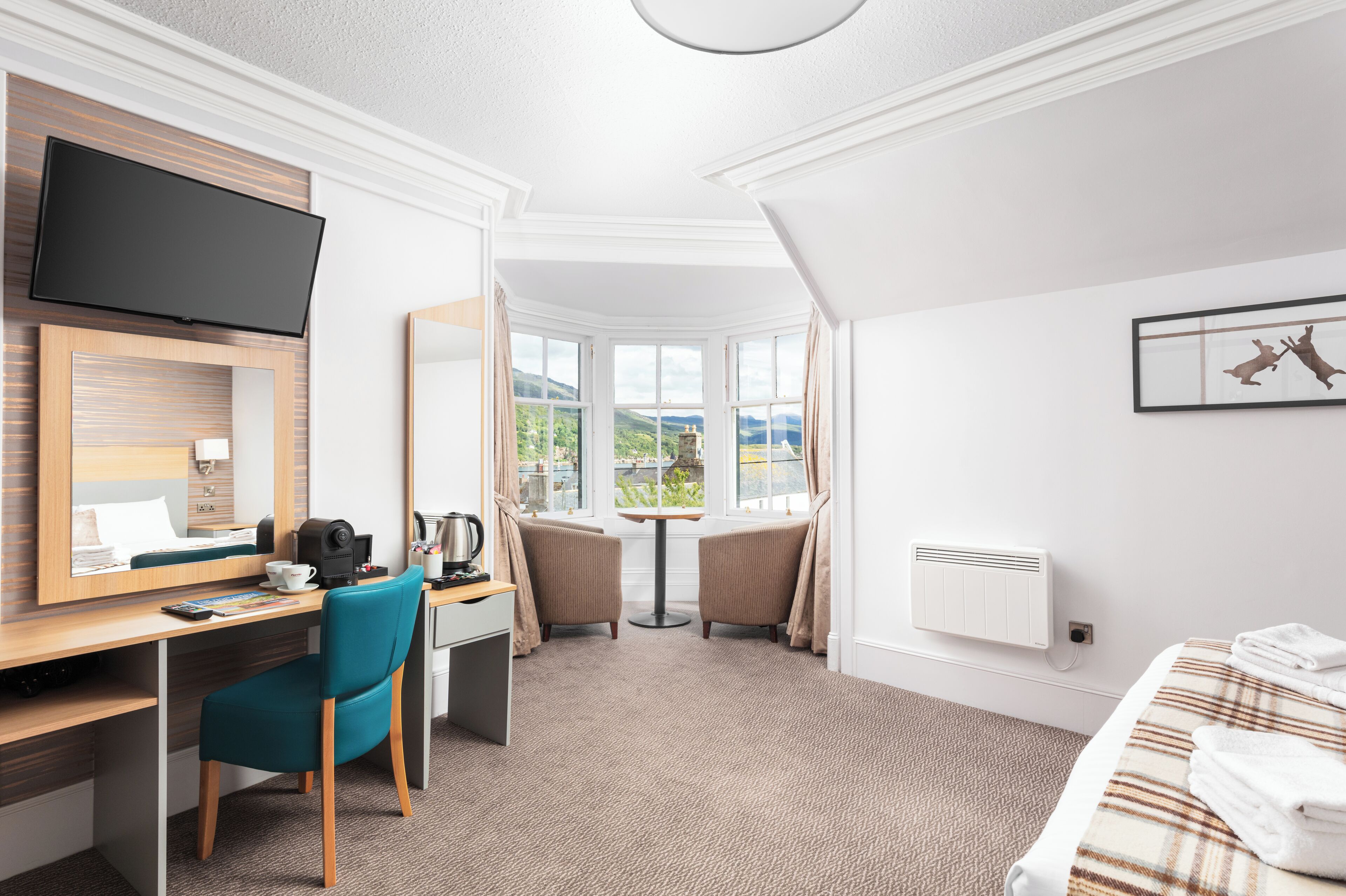 A hotel room with a desk, chair, bed, TV, window, and artwork.