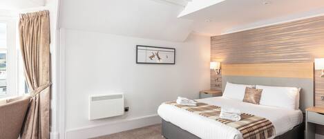 A modern hotel room with a large bed, a bedside table, a chair, a radiator, and a window with curtains.
