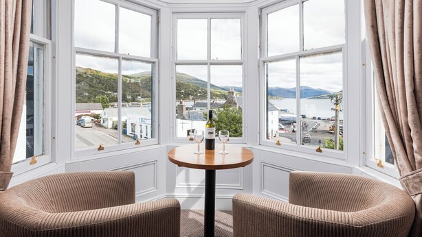 A corner room with a bay window overlooking a town and hills. There is a round table with two chairs and a bottle of wine. - Caledonian Hotel (Ullapool)