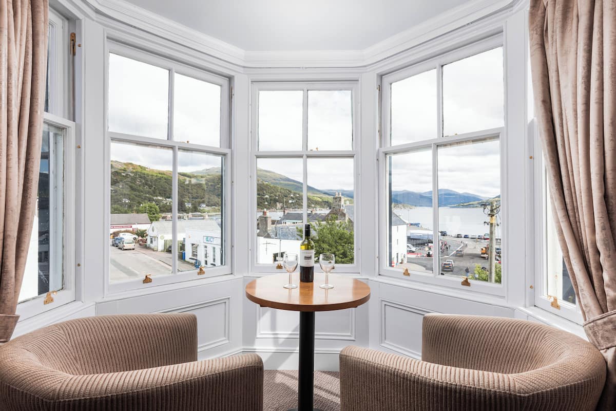 A corner room with a bay window overlooking a town and hills. There is a round table with two chairs and a bottle of wine.