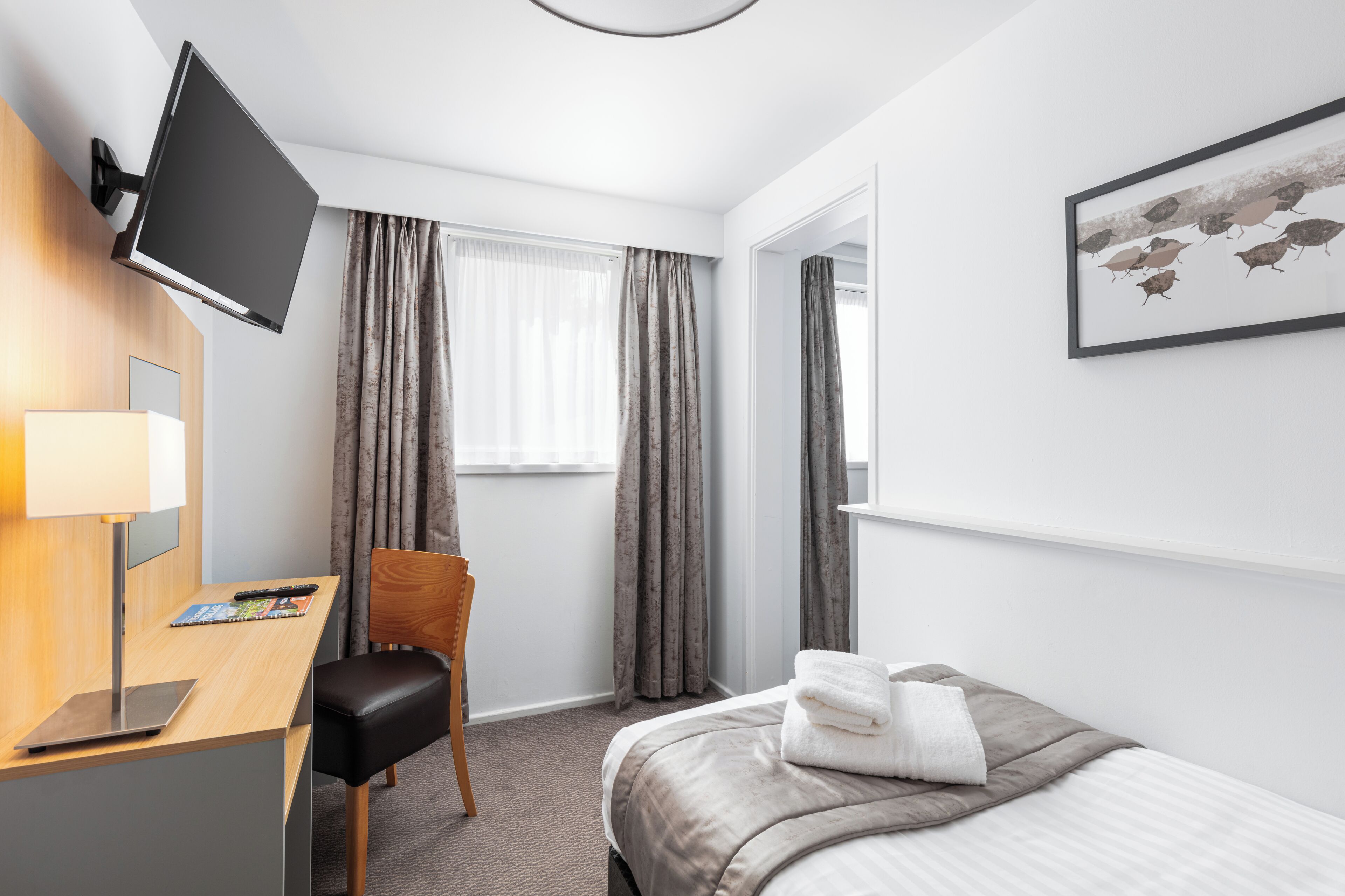 A hotel room with a bed, a desk with a laptop, a chair, a TV mounted on the wall, and a window with curtains.