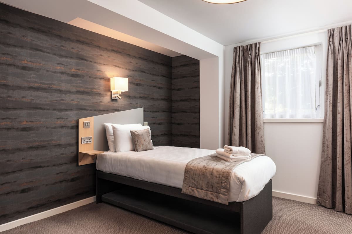 A modern hotel room with a bed, a nightstand, a wall-mounted lamp, and a window with curtains.