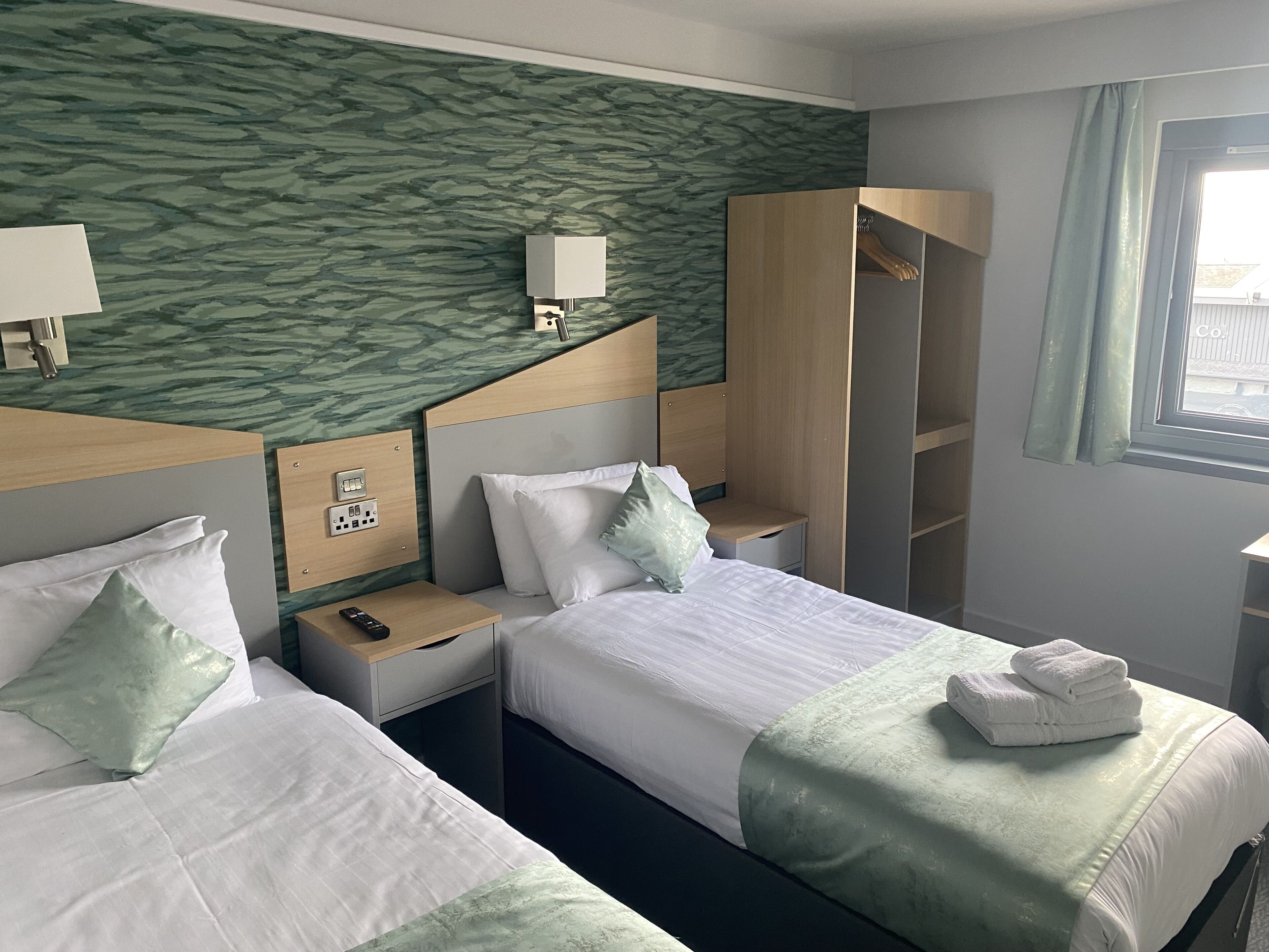 A hotel room with two beds, a wardrobe, a nightstand with a lamp, and a window.