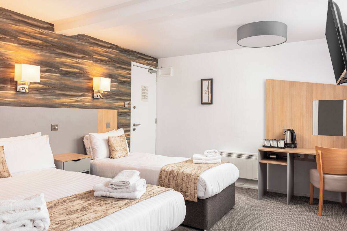 A hotel room with two beds, a desk, a chair, a flat-screen TV, and a wall-mounted lamp.