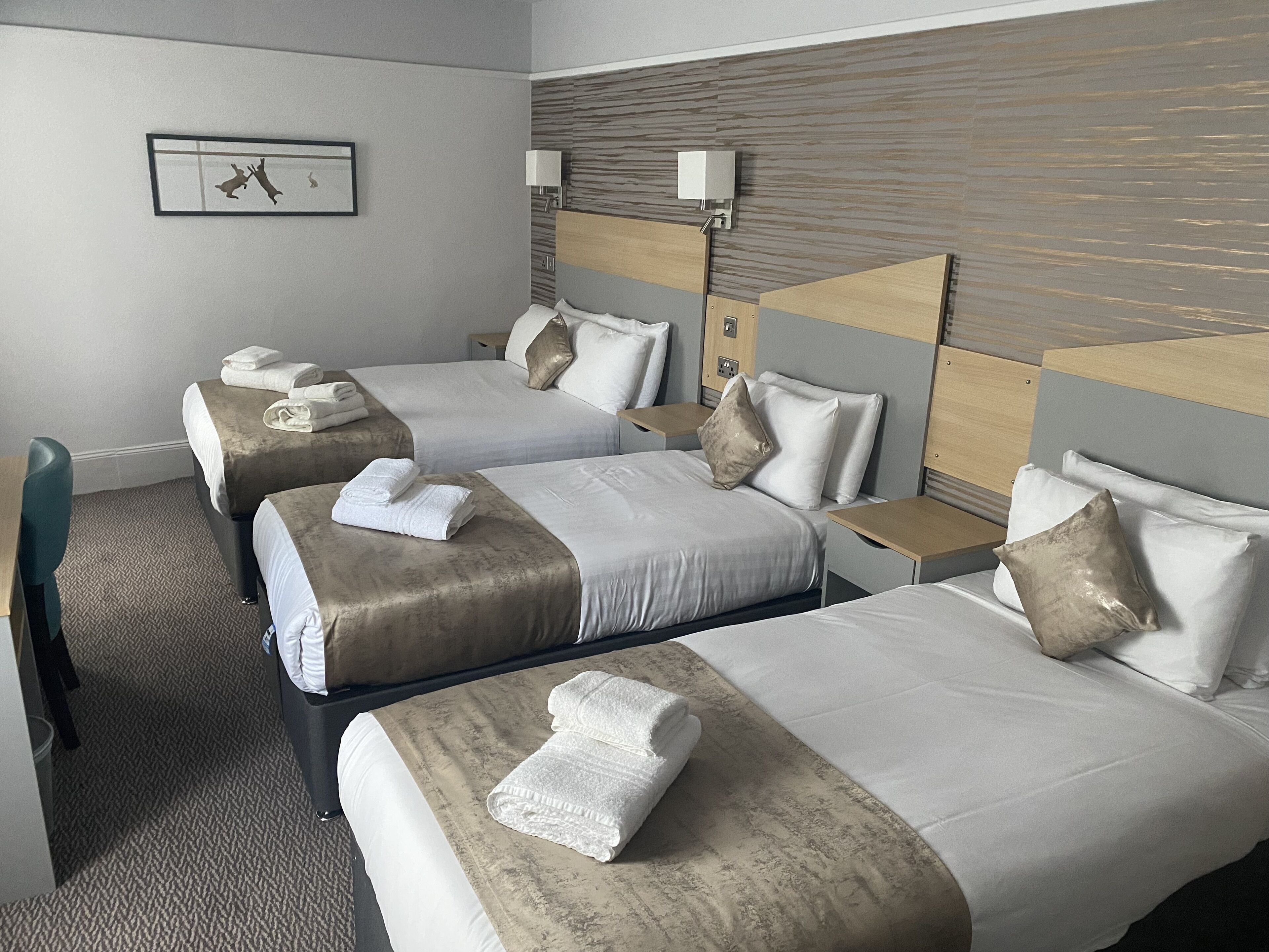 A hotel room with two beds, each with a pillow and a folded towel. There is a small table and a chair, a wall-mounted lamp, and a framed picture on the wall.
