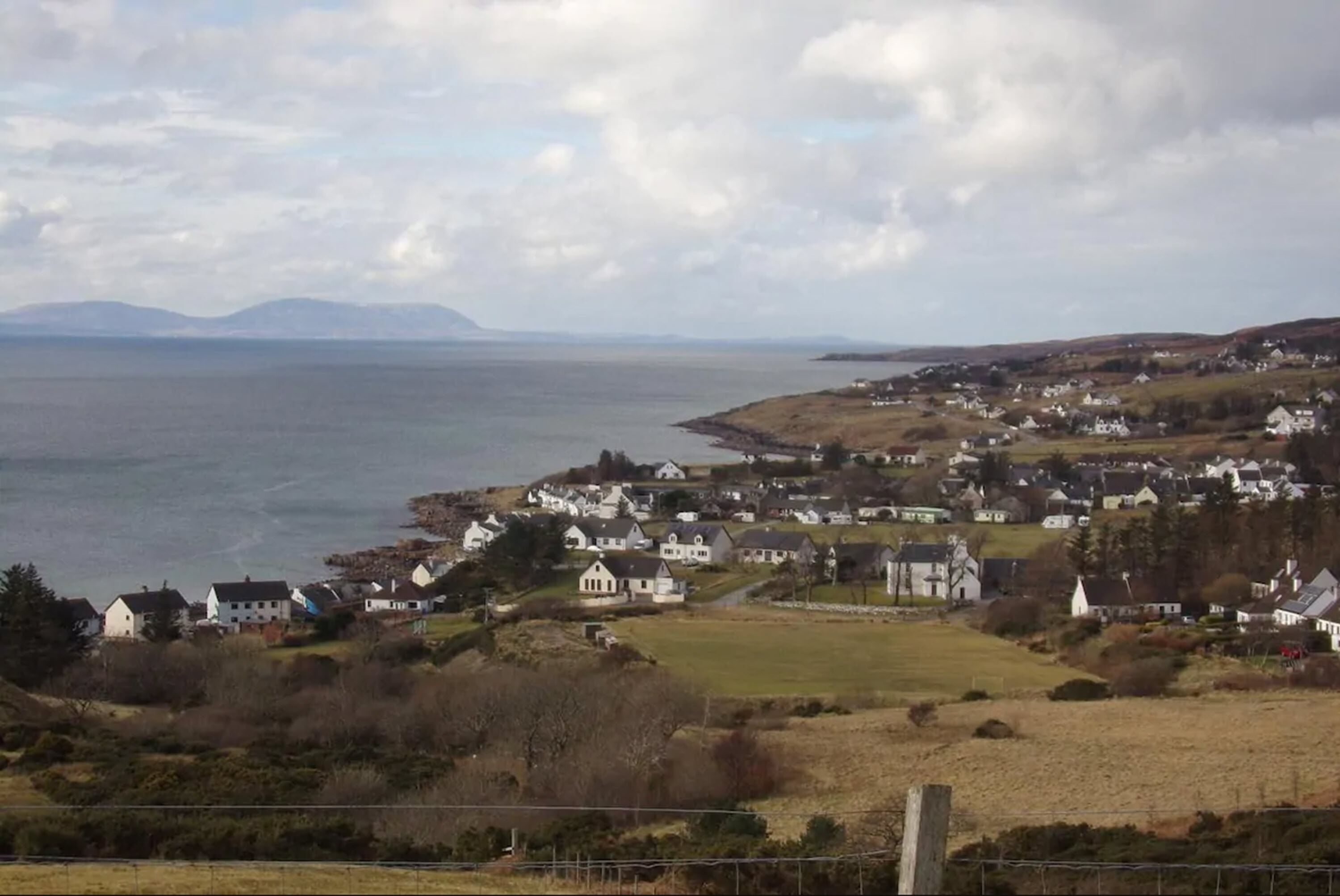 A coastal village with houses and a church, surrounded by hills and a body of water.