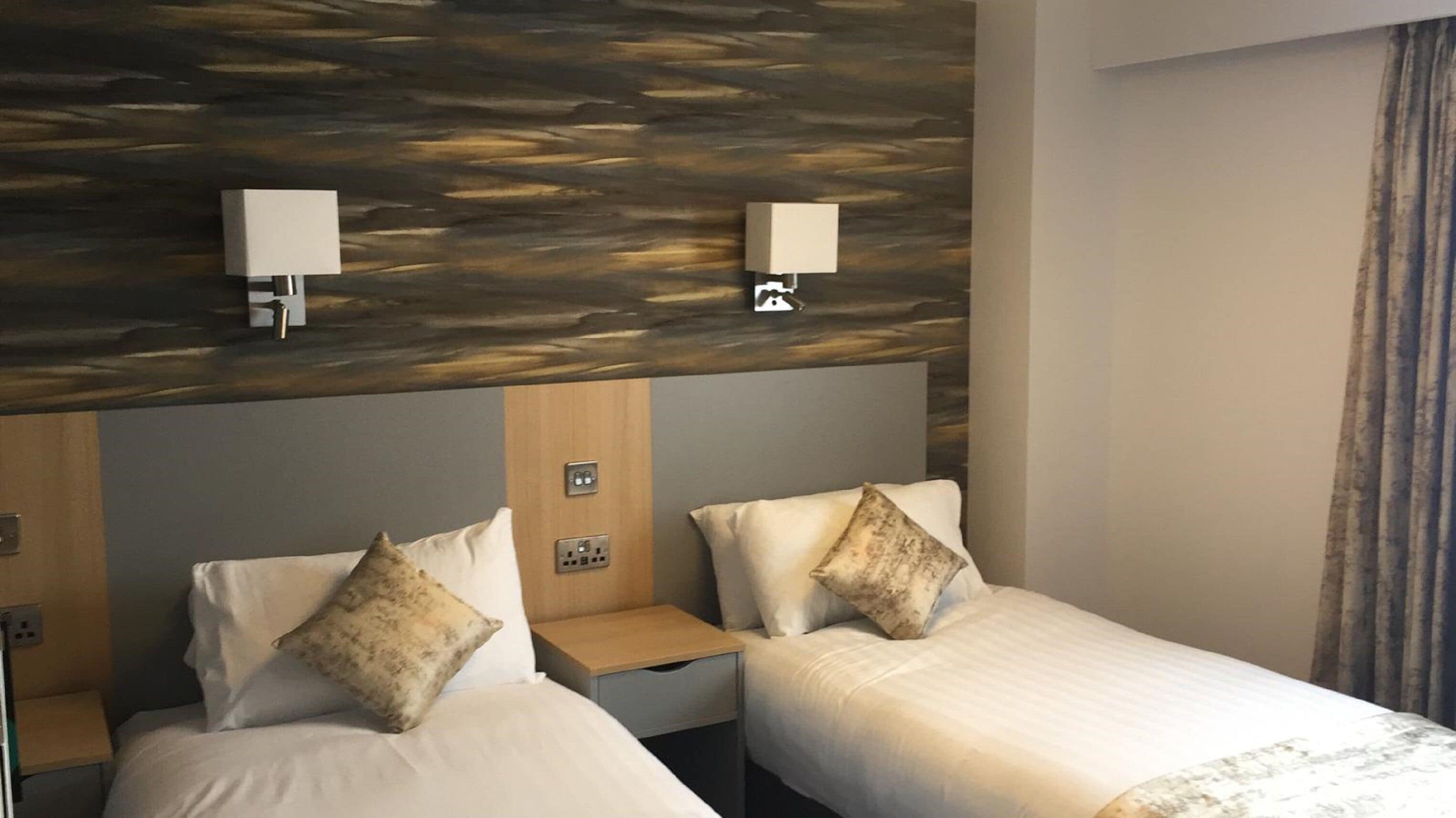 A hotel room with two beds, each with a pillow and a nightstand. There are wall-mounted lamps above the beds and a power outlet on the wall.