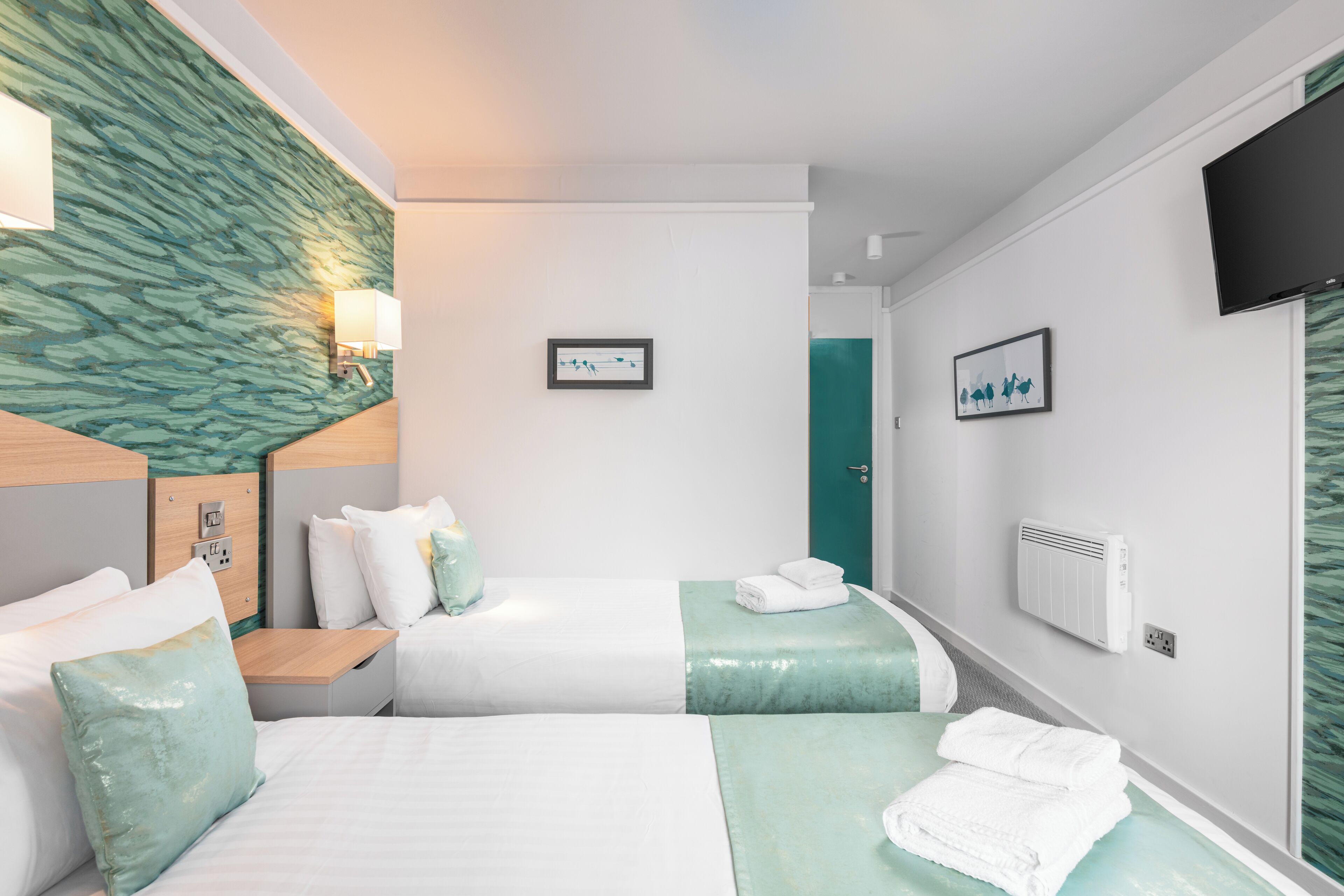 A hotel room with two beds, a wall-mounted TV, a power outlet, and a door.
