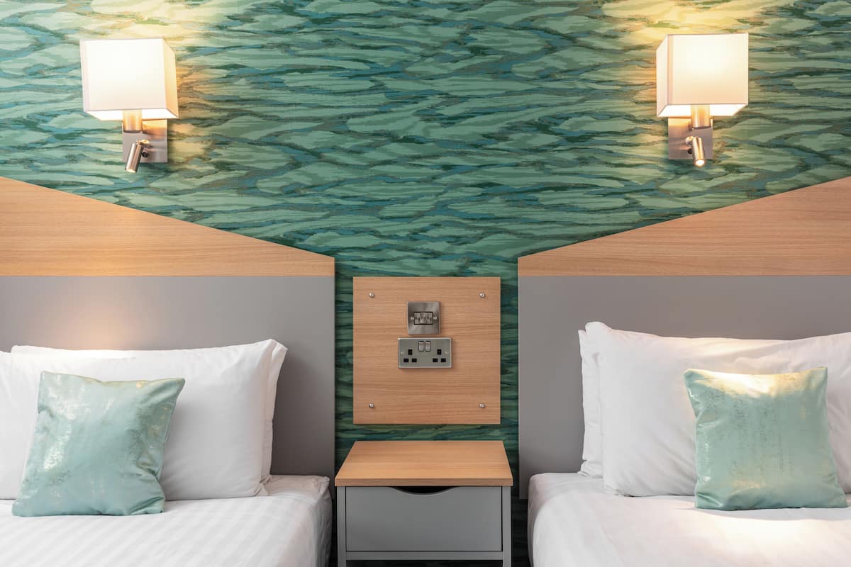 A hotel room with two beds, a nightstand, a wall-mounted electrical outlet, and two wall-mounted lamps.
