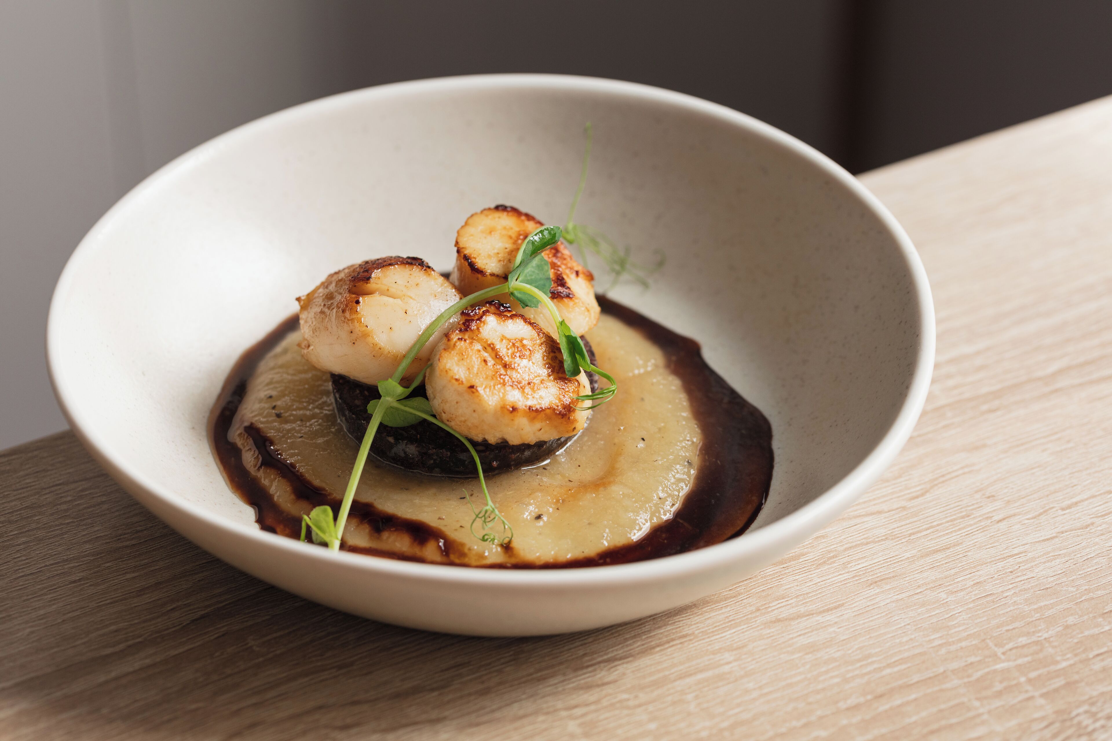 A white bowl with seared scallops and a dark sauce, placed on a wooden surface.