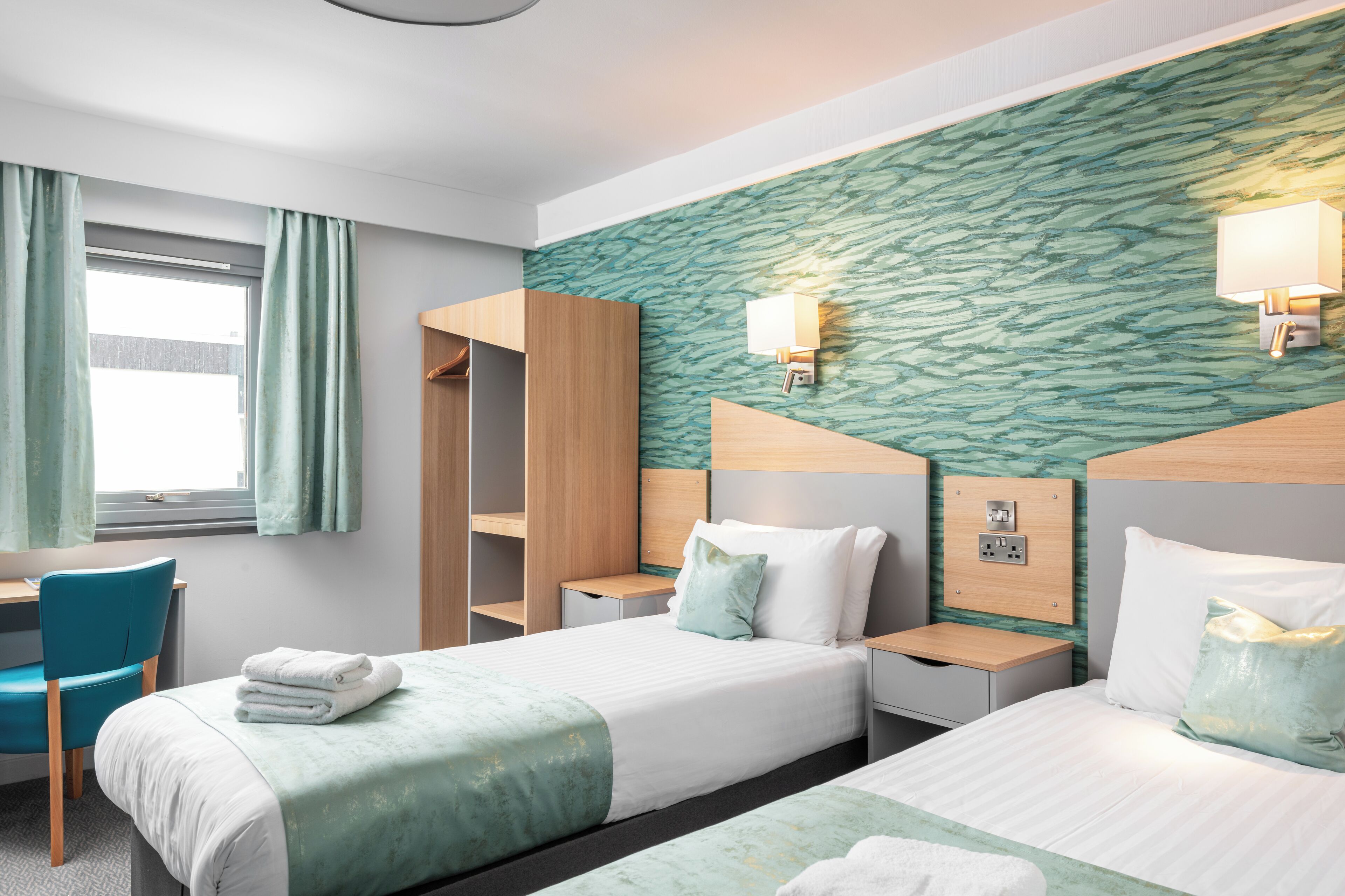 A hotel room with two beds, a desk, and a chair. There are wall-mounted lamps and a window with curtains.