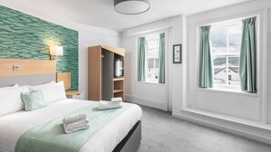 A hotel room with a bed, a bedside table, a wardrobe, a window with curtains, and a view of buildings outside. - Caledonian Hotel (Ullapool)