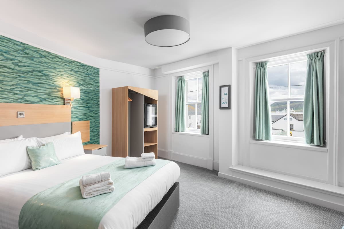 A hotel room with a bed, a bedside table, a wardrobe, a window with curtains, and a view of buildings outside.