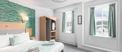 A hotel room with a bed, a bedside table, a wardrobe, a window with curtains, and a view of buildings outside.