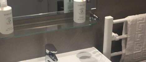 Un baño con un lavamanos blanco, una repisa de vidrio con artículos de aseo, un perchero para toallas y una obra de arte enmarcada en la pared.