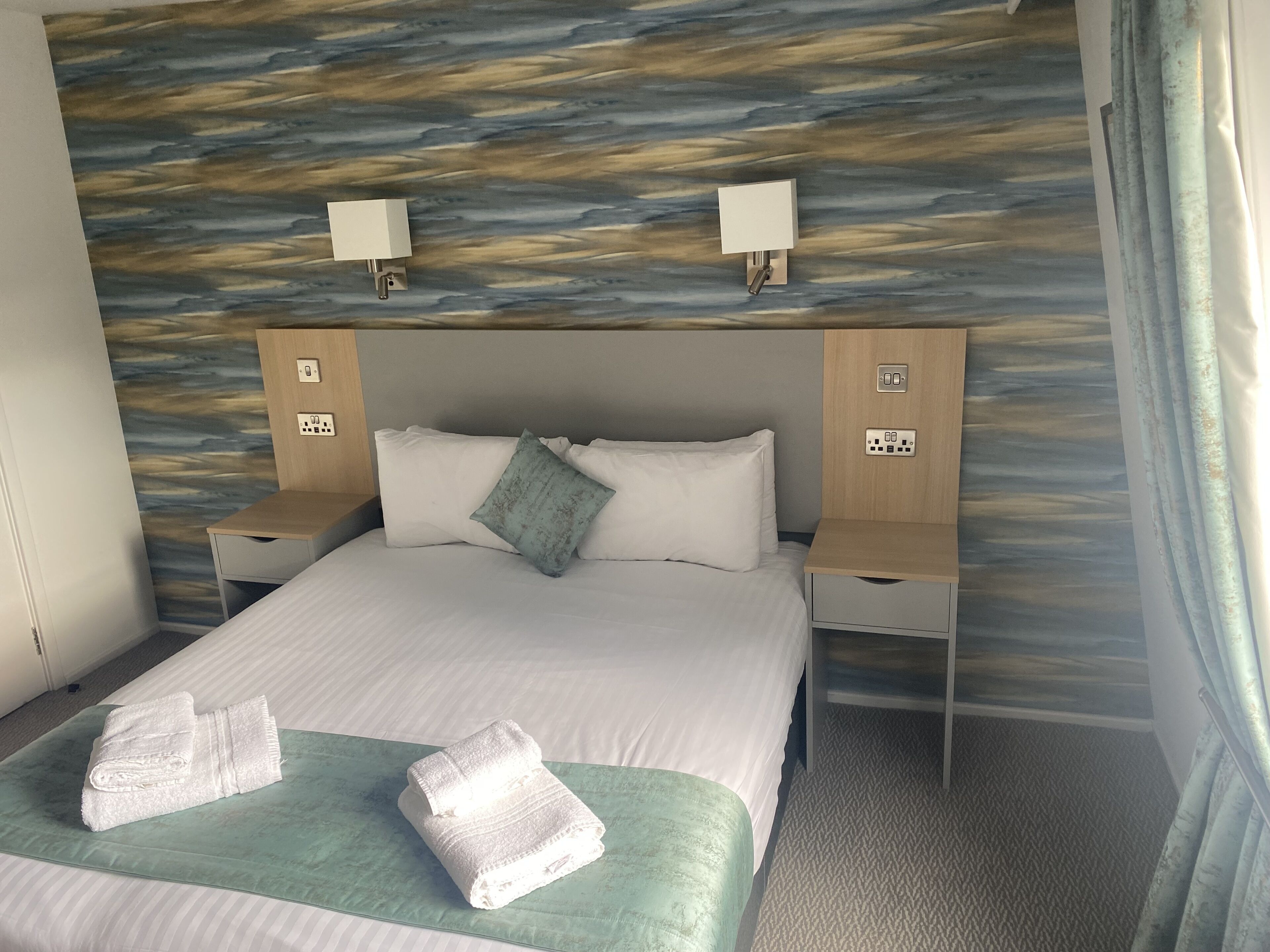 Habitación de hotel con una cama grande, dos mesitas de noche, una lámpara de pared y un enchufe en la cabecera.