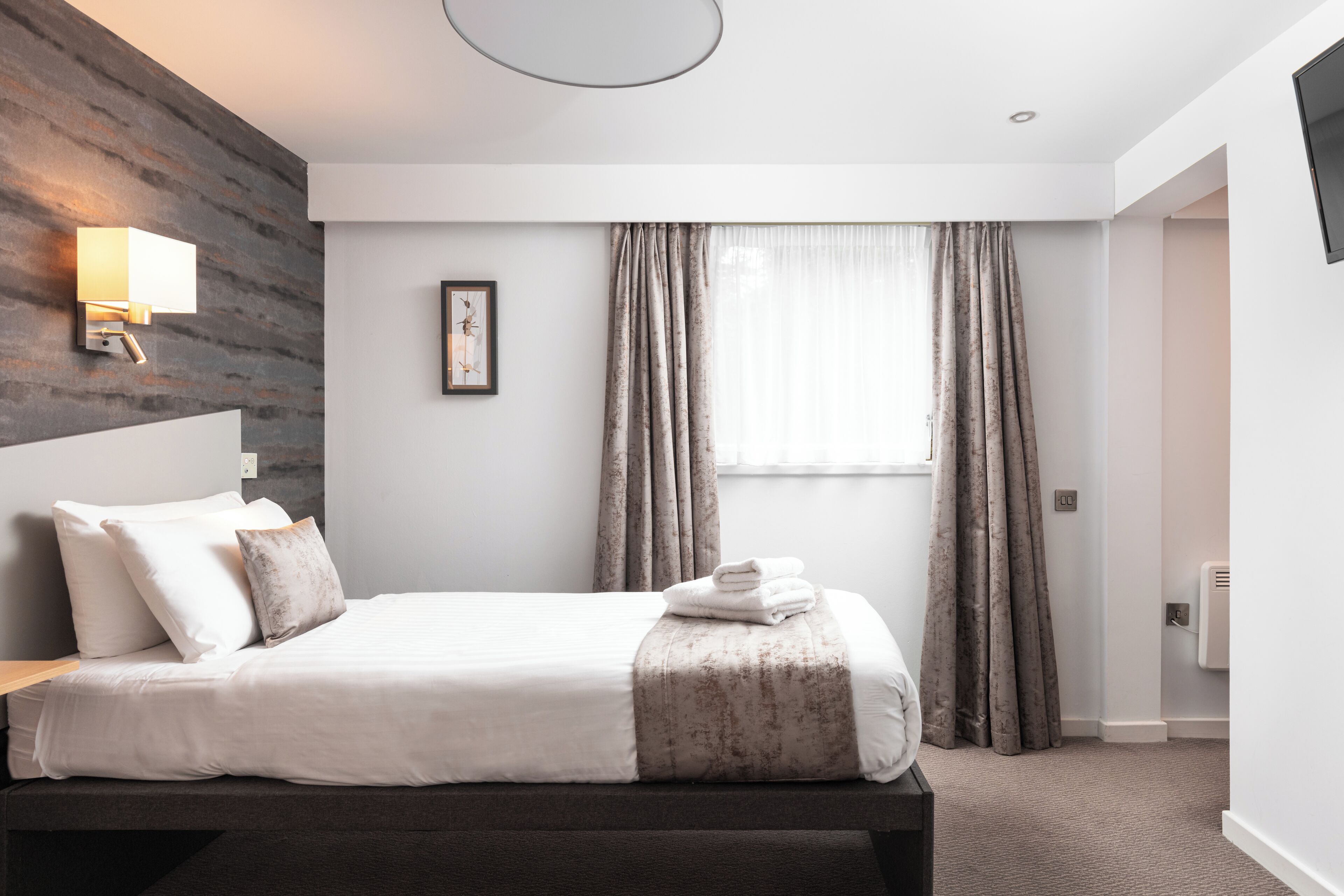 Una habitación de hotel moderna con una cama grande, una mesita de noche, una lámpara de pared, un cuadro enmarcado y una ventana con cortinas.