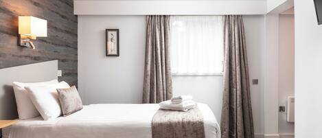 Una habitación de hotel moderna con una cama grande, una mesita de noche, una lámpara de pared, un cuadro enmarcado y una ventana con cortinas.