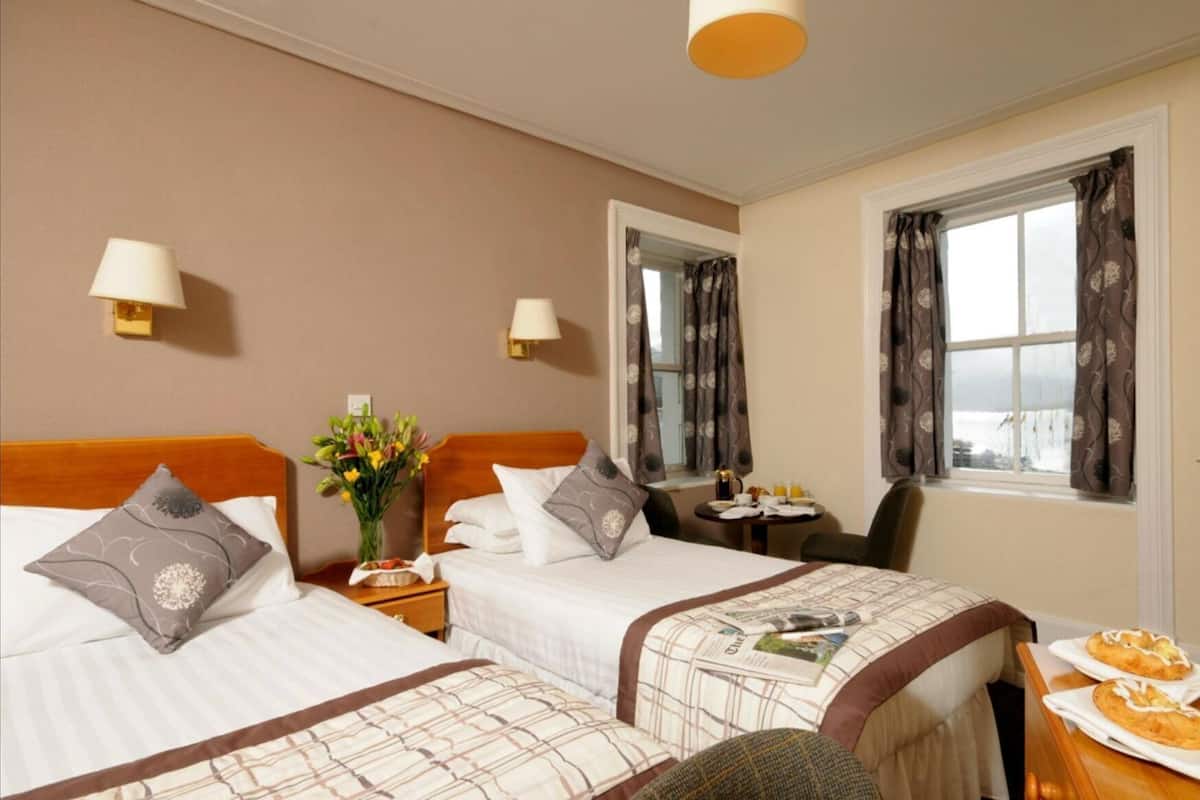 A hotel room with two beds, a small table with breakfast items, a chair, and a window with curtains.