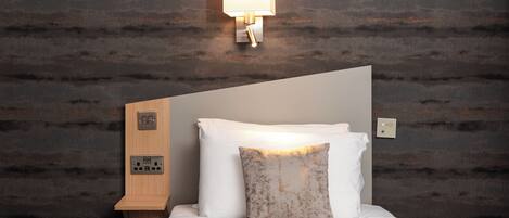 A neatly made bed with a white pillow and a textured throw pillow, a wooden bedside table with a power outlet, and a wall-mounted light fixture.