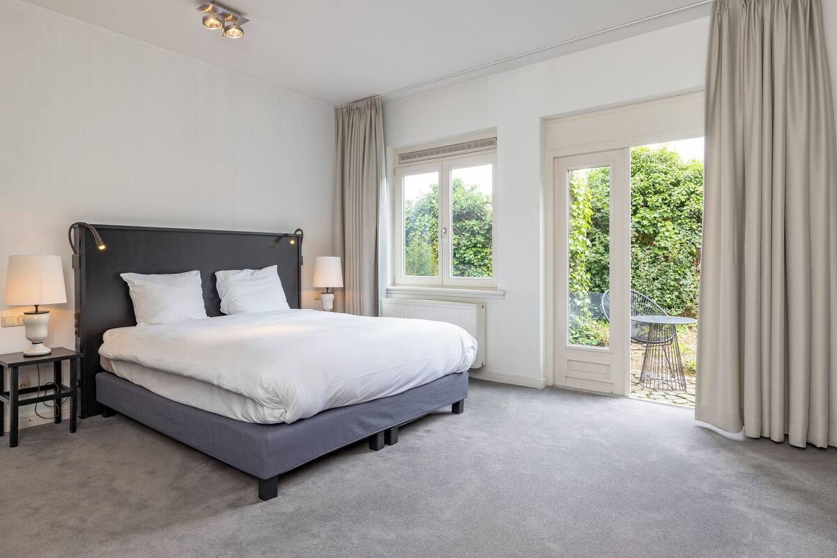 A modern bedroom with a large bed, bedside lamps, and a view of the outdoors through a sliding glass door.