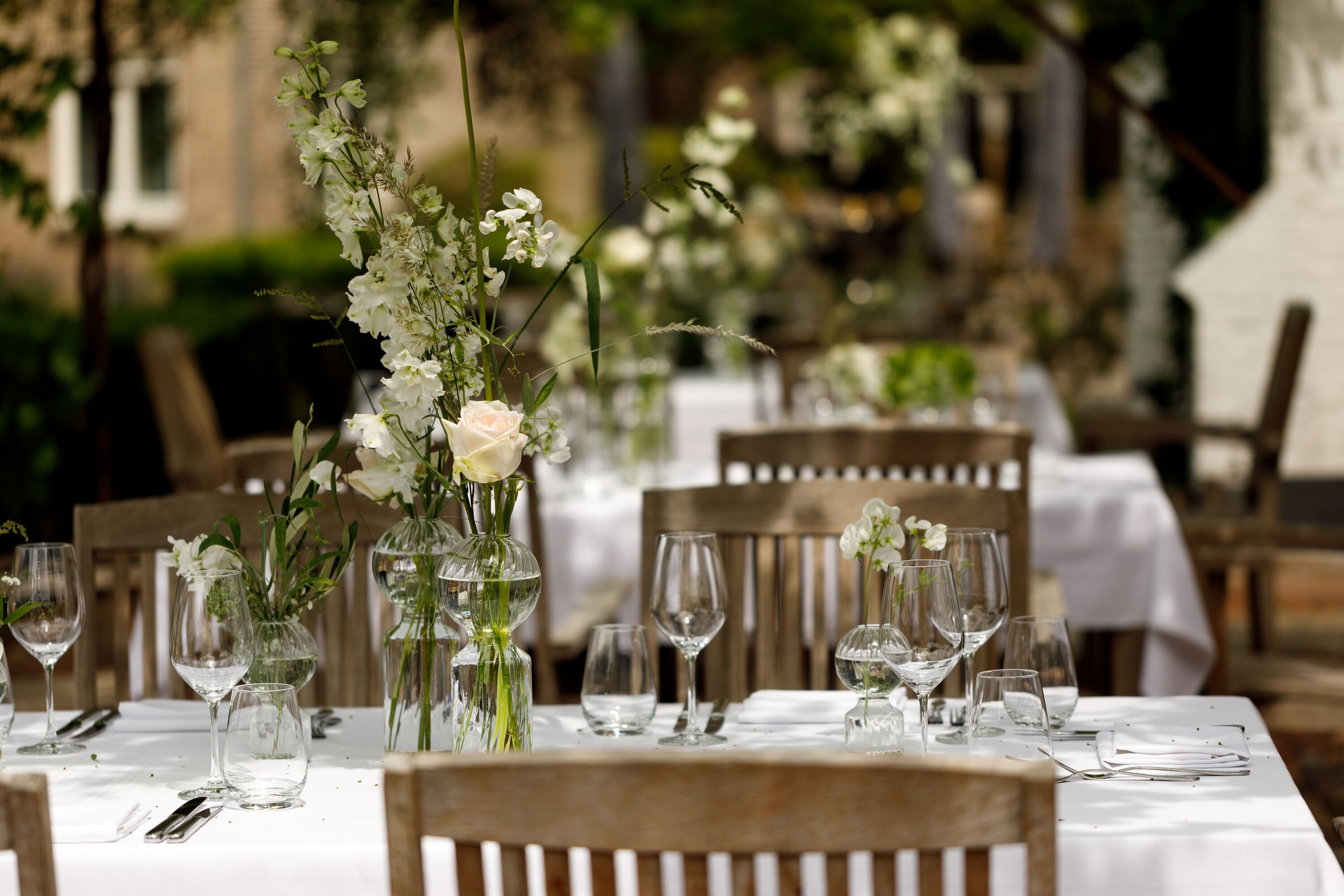 Ein Essplatz draußen mit weißen Tischdecken, klarem Glas und floralen Mittelstücken.