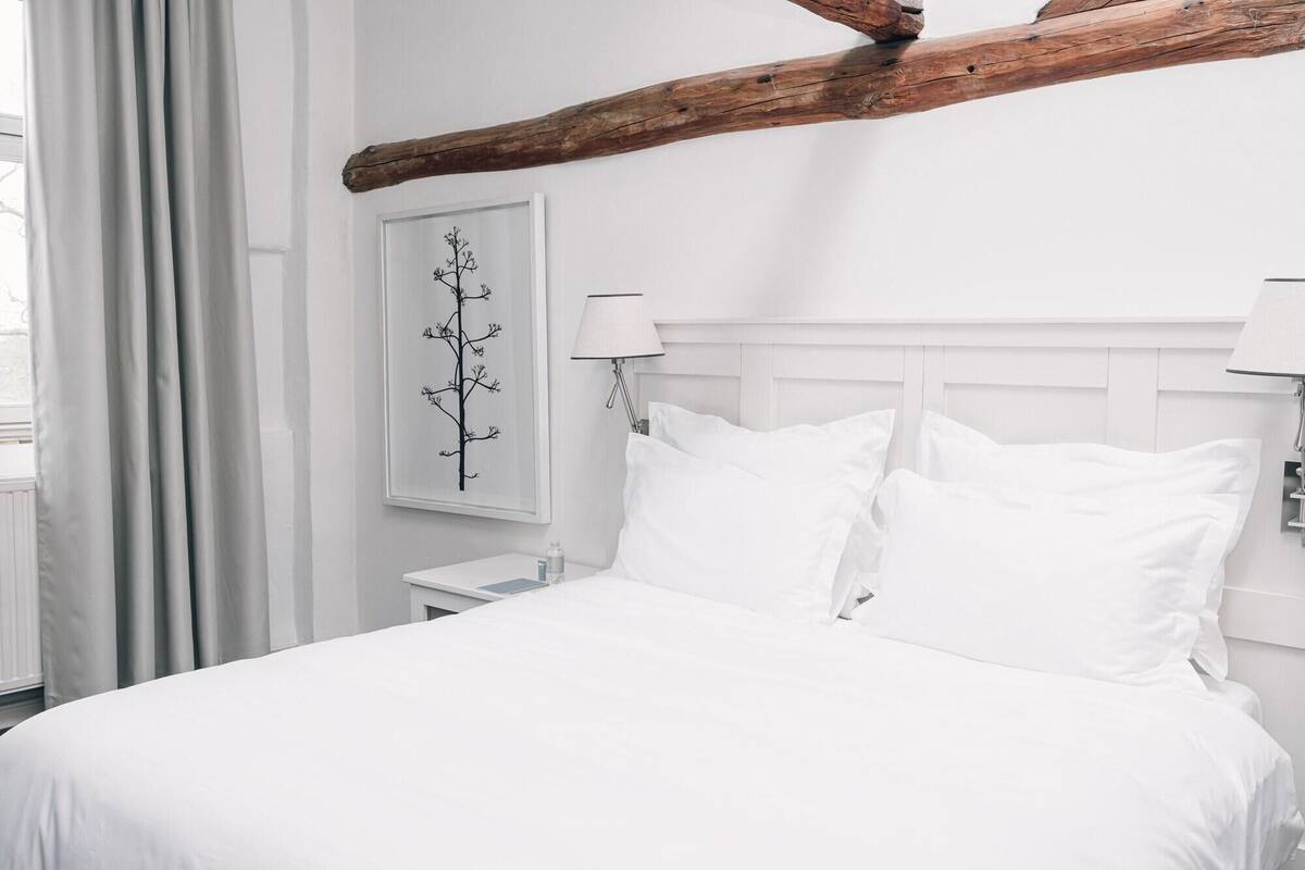 A white bed with two pillows, a nightstand with a lamp, a wall-mounted picture, and a wooden beam above the bed.