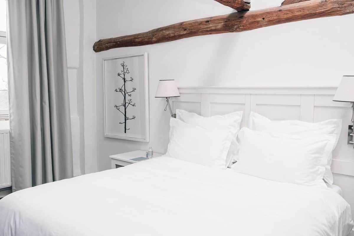 Un lit blanc avec deux oreillers, une table de chevet avec une lampe, un tableau accroché au mur et une poutre en bois au-dessus du lit.