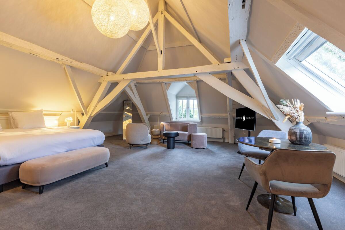 A spacious room with a high ceiling, a large bed, a seating area with a table, and a window providing natural light.