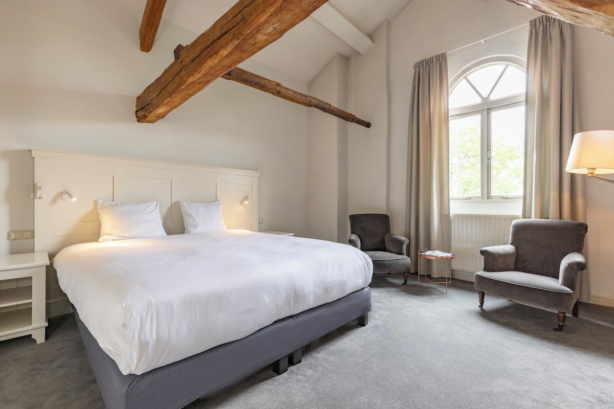 A bedroom with a large bed, a chair, a nightstand, and a window with curtains.