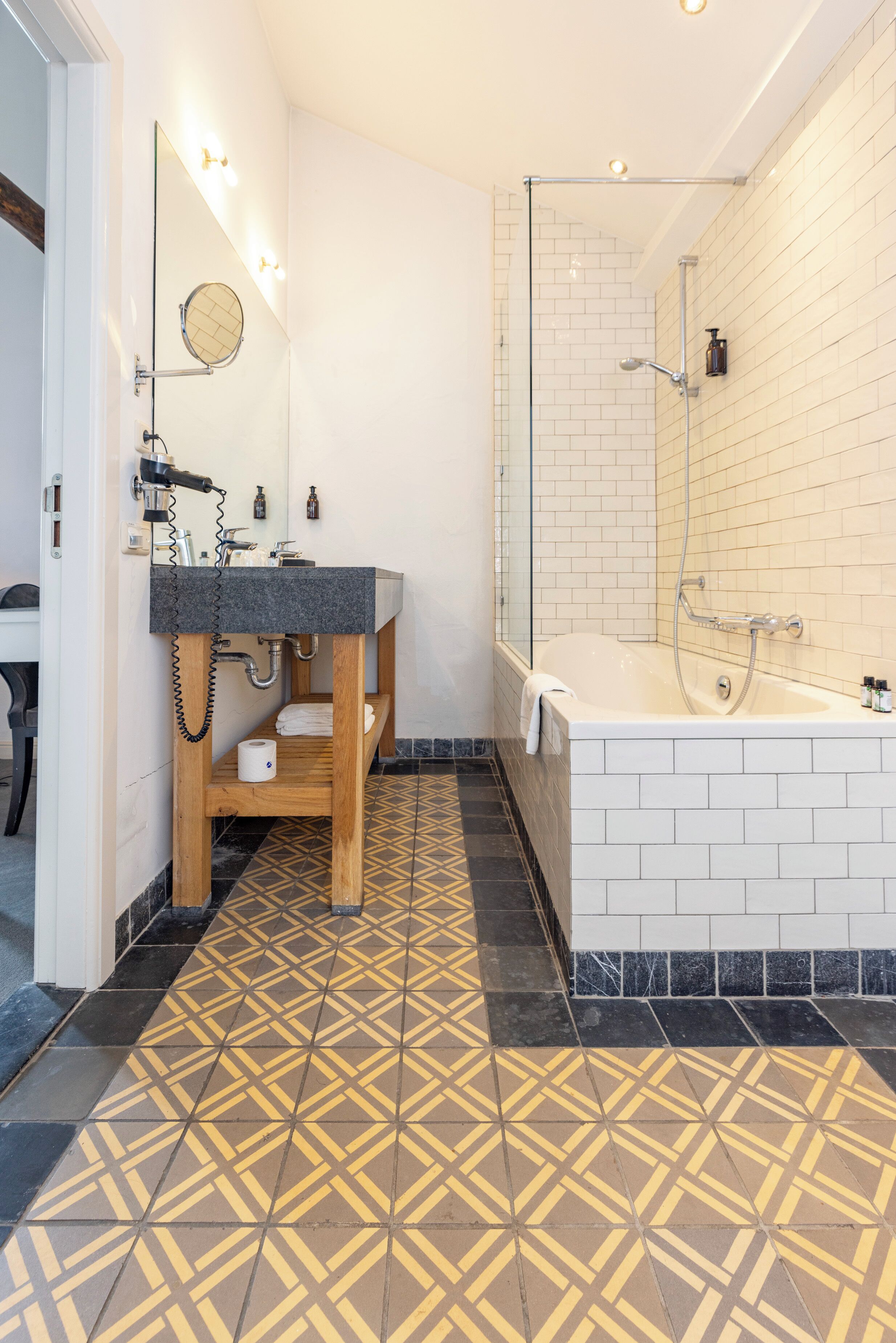 A modern bathroom with a wooden vanity, a large mirror, a bathtub, and patterned floor tiles.