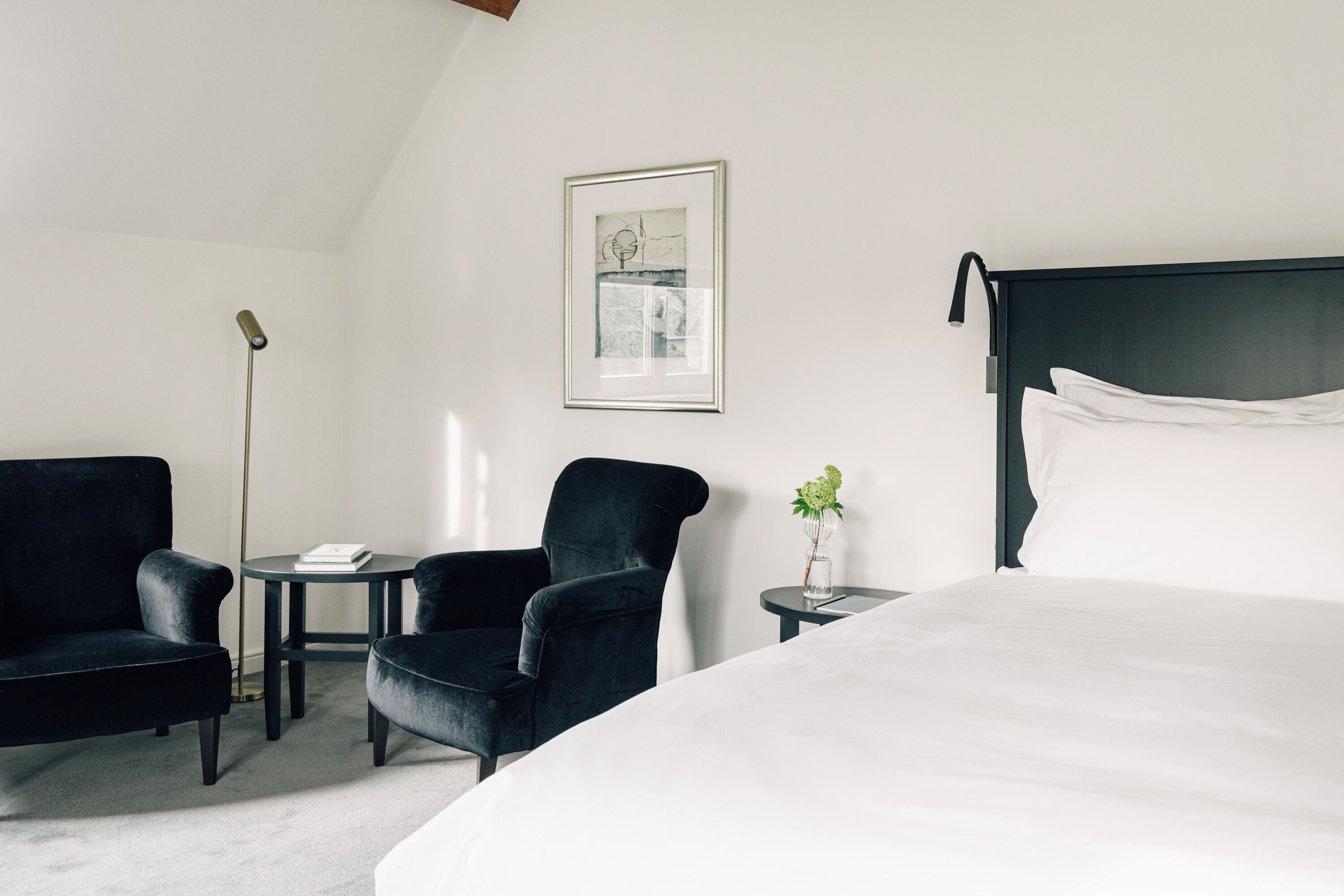 Quarto de hotel com uma cama grande, duas poltronas pretas, uma mesa redonda pequena, um abajur, um quadro na parede e um vaso com flores sobre uma mesinha lateral.
