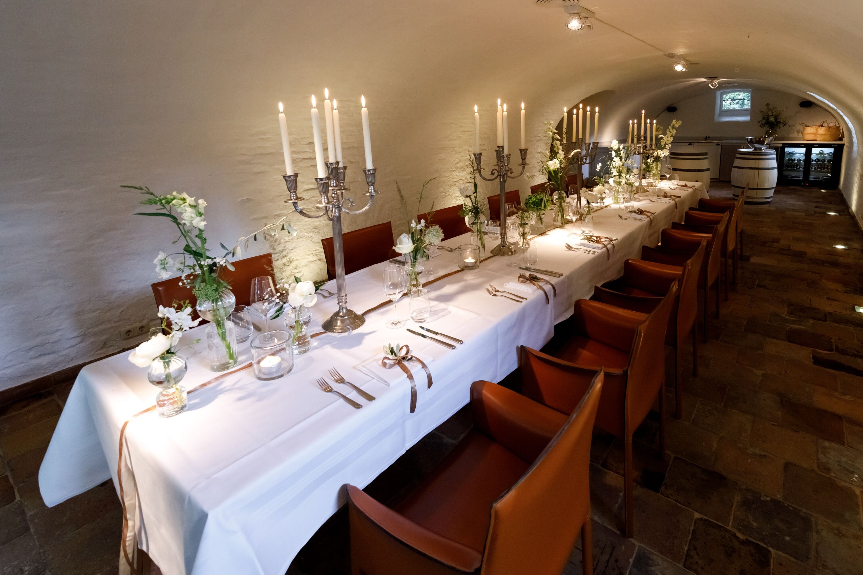 A long dining table set with candles, flowers, and fine china in a stone-walled room with a vaulted ceiling.