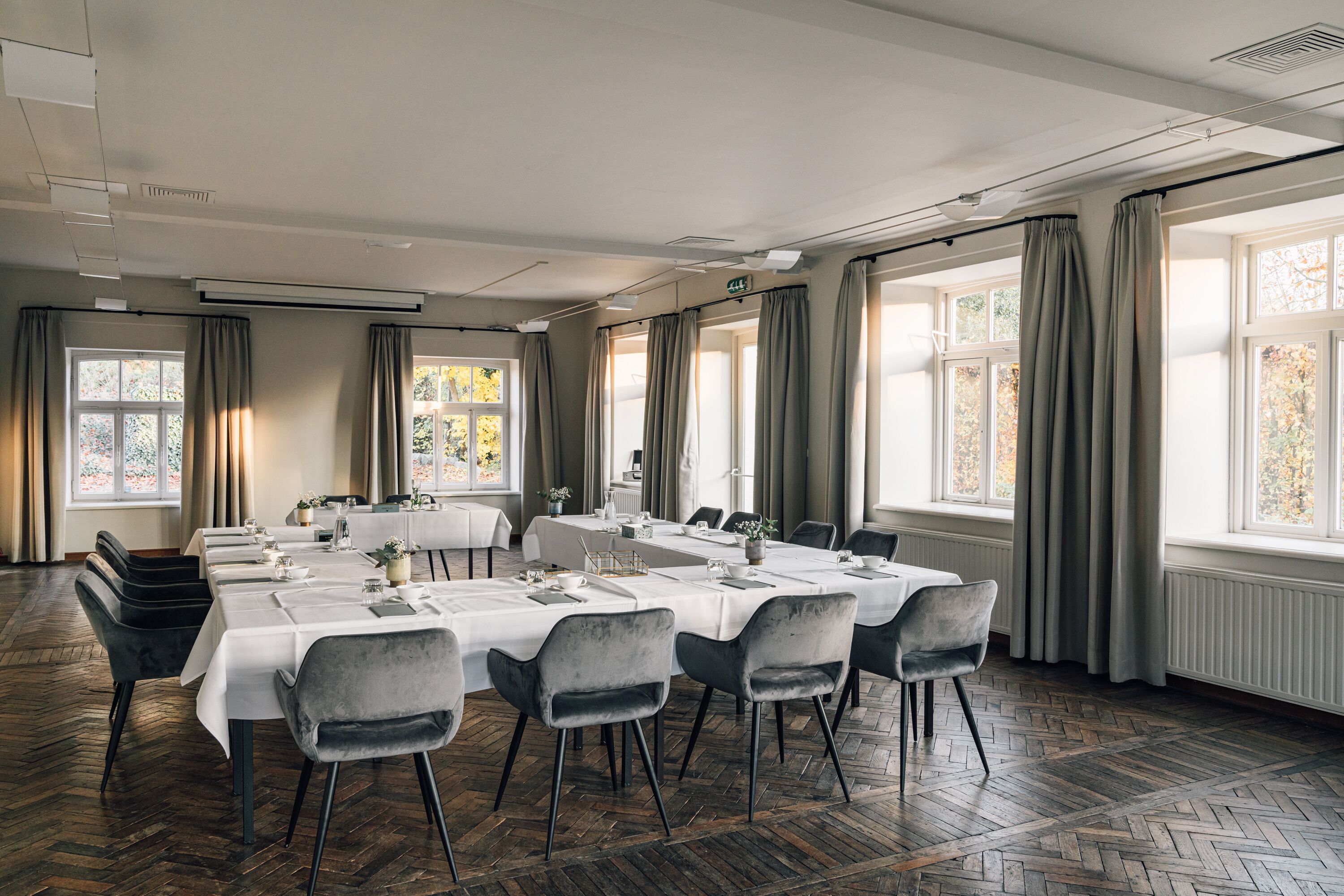 Un salón de conferencias con una larga mesa dispuesta para una reunión, amplias ventanas con cortinas y un piso de madera con diseño de espiga.
