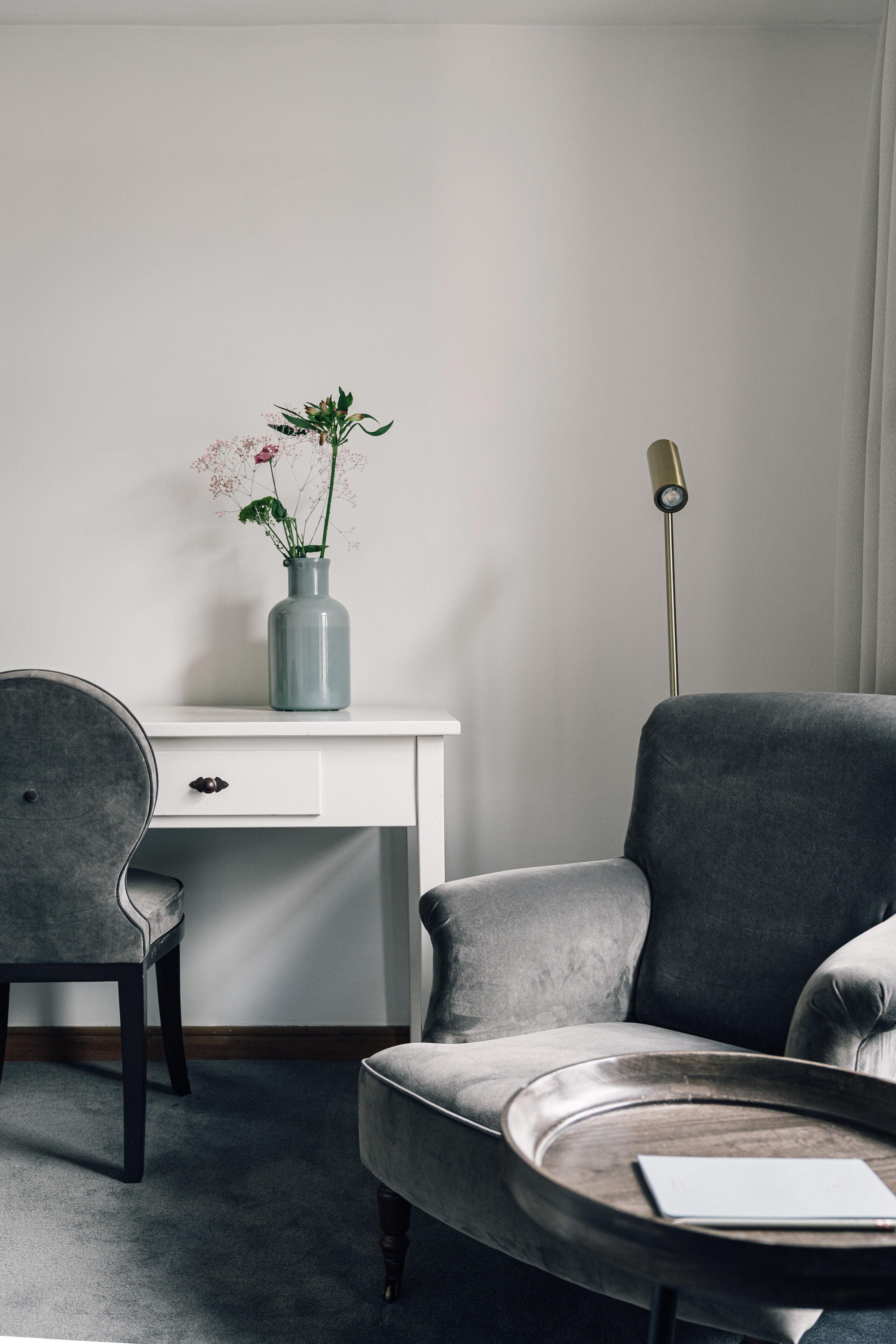 Una habitación moderna con un sillón gris, un escritorio blanco y un florero con flores.