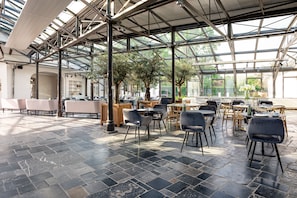 A spacious indoor area with a high ceiling and large windows, featuring multiple seating arrangements and a central plant. - Fletcher Hotel Château De Raay (Baarlo)
