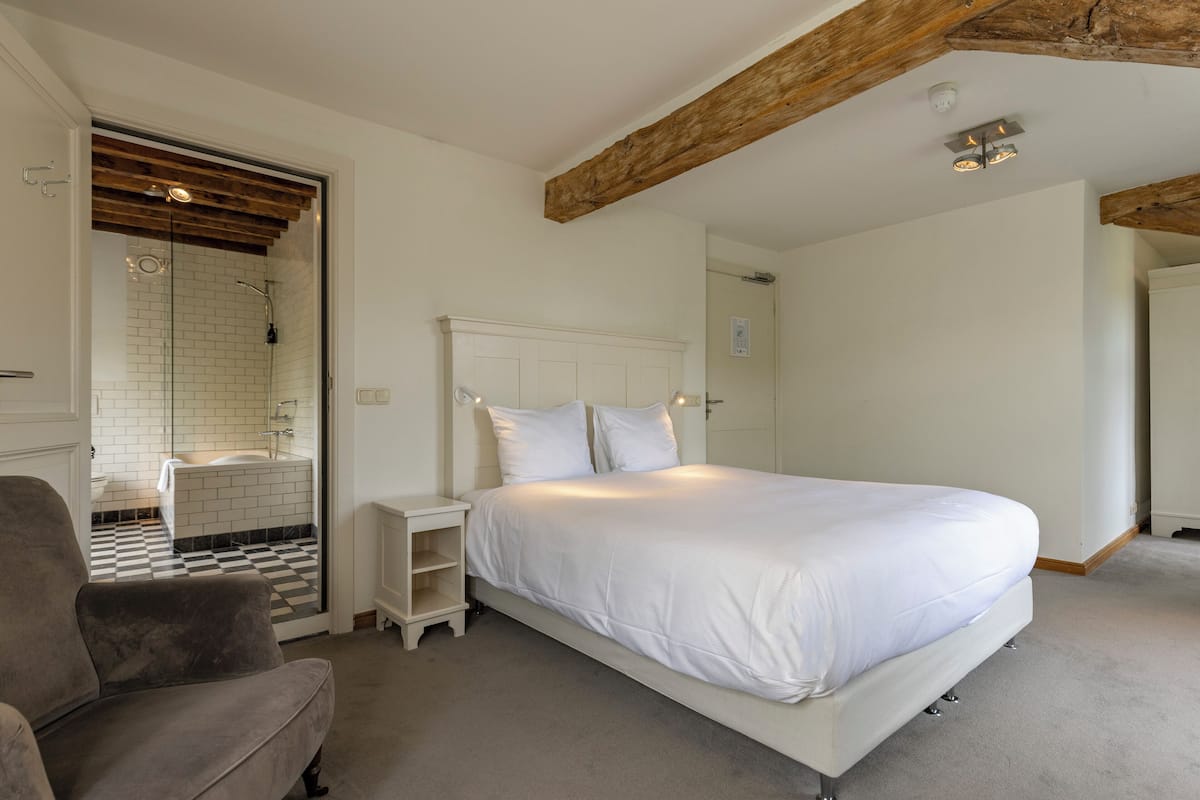 A bedroom with a large bed, a nightstand, a chair, and a bathroom with a shower.
