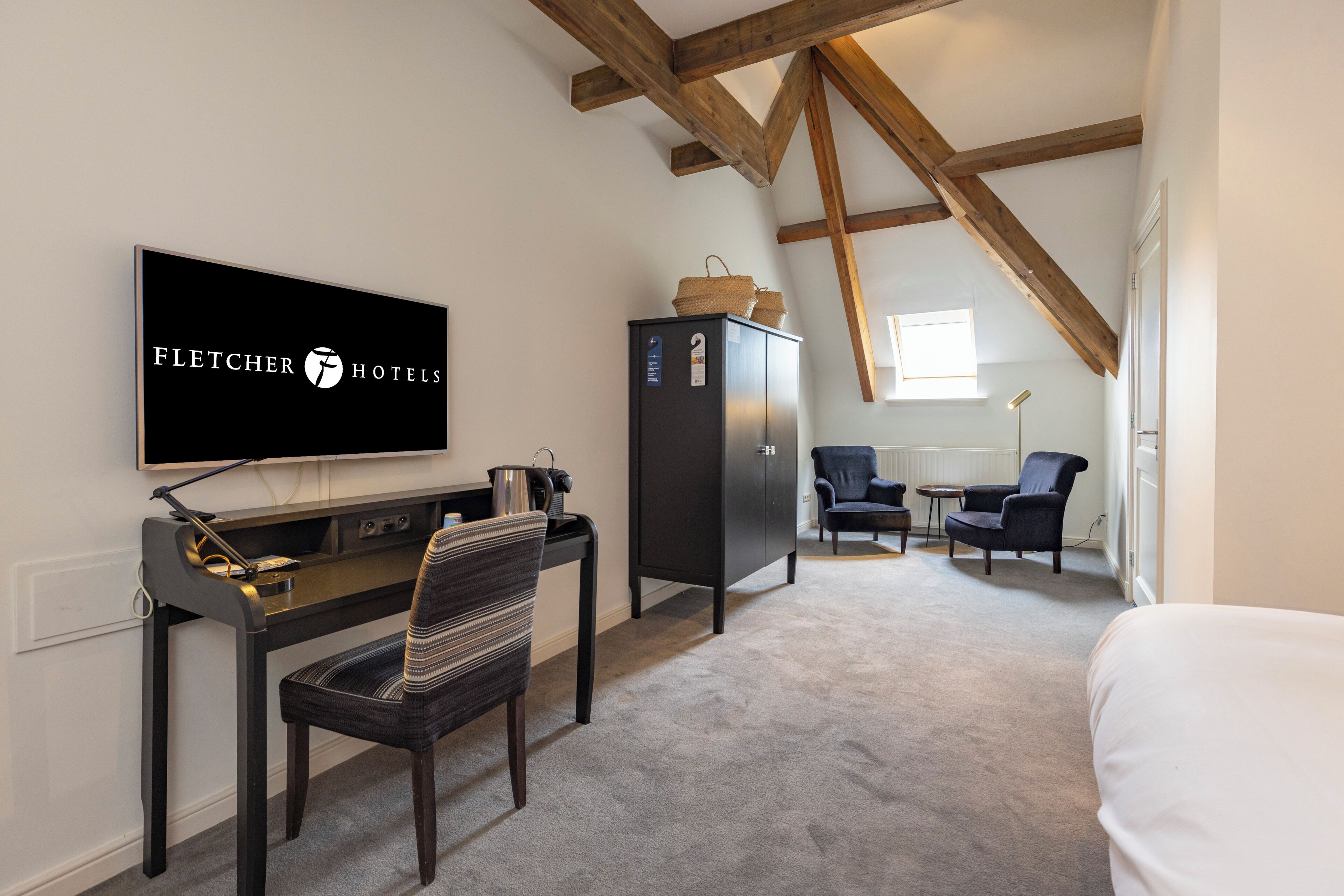 Ein Hotelzimmer mit Schreibtisch, Stuhl, Fernseher, Kleiderschrank und zwei Sesseln.