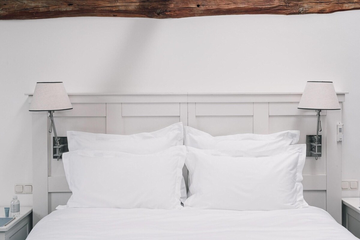 Een wit bed met twee kussens, een houten balk in het plafond en twee bedlampjes.