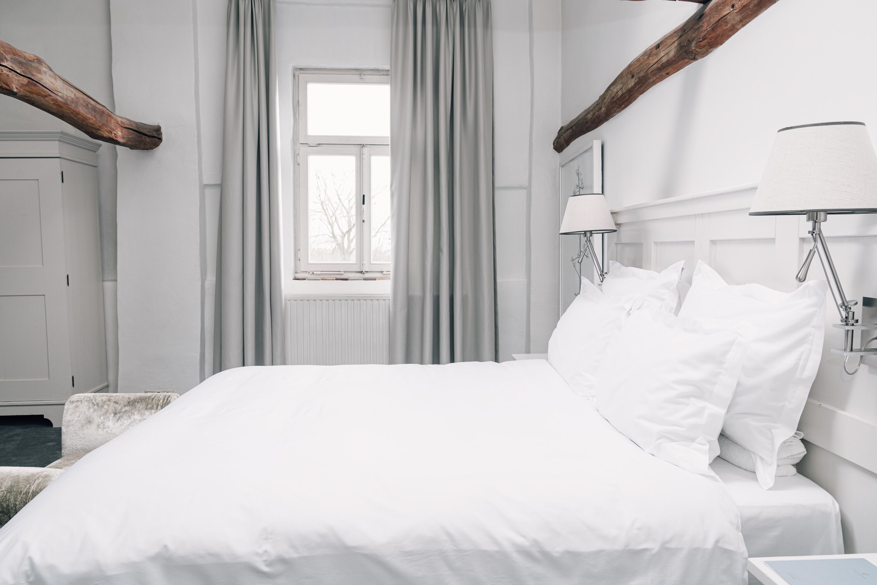 A white bed with multiple pillows, a bedside table, and two wall-mounted lamps. A window with curtains is visible in the background.