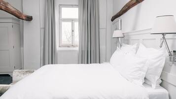 A white bed with multiple pillows, a bedside table, and two wall-mounted lamps. A window with curtains is visible in the background.