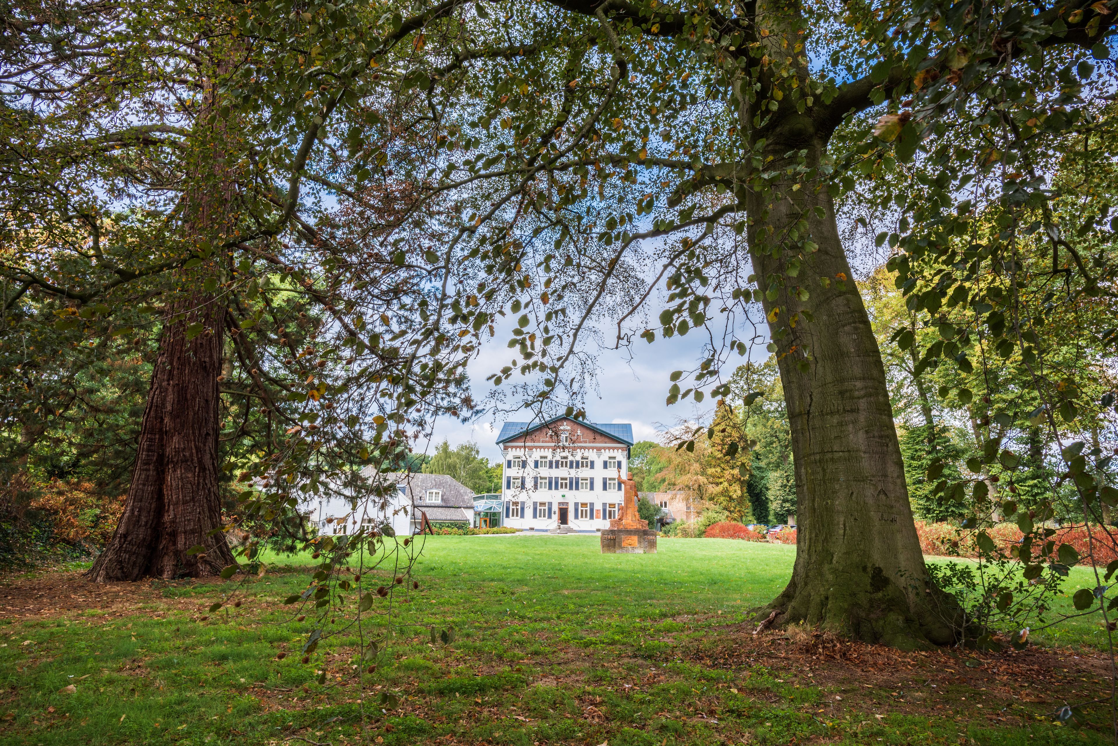 Een groot gebouw met een symmetrische gevel, omringd door een verzorgd gazon en volwassen bomen.