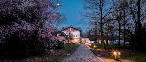 A cobblestone pathway leading to a historic building with a traditional architectural style, surrounded by flowering trees and illuminated by street lamps.