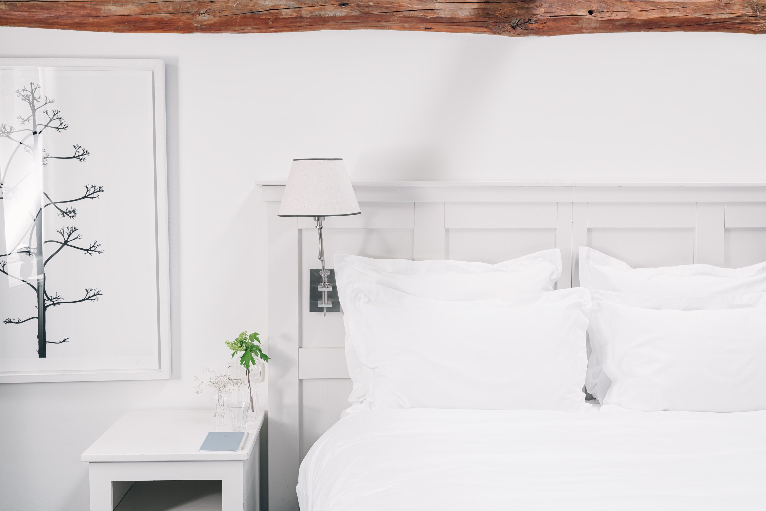 Un dormitorio blanco con una cama, una mesita de noche, una lámpara y una obra de arte en la pared.