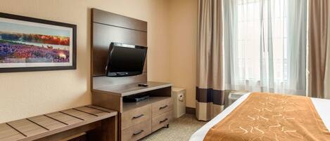 A hotel room with a bed, a TV on a wooden stand, a small desk with drawers, a picture on the wall, and a window with curtains.