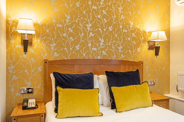 Habitación de hotel con cabecera de madera, dos lámparas de noche, un teléfono y una cama con almohadas amarillas y azul marino.
