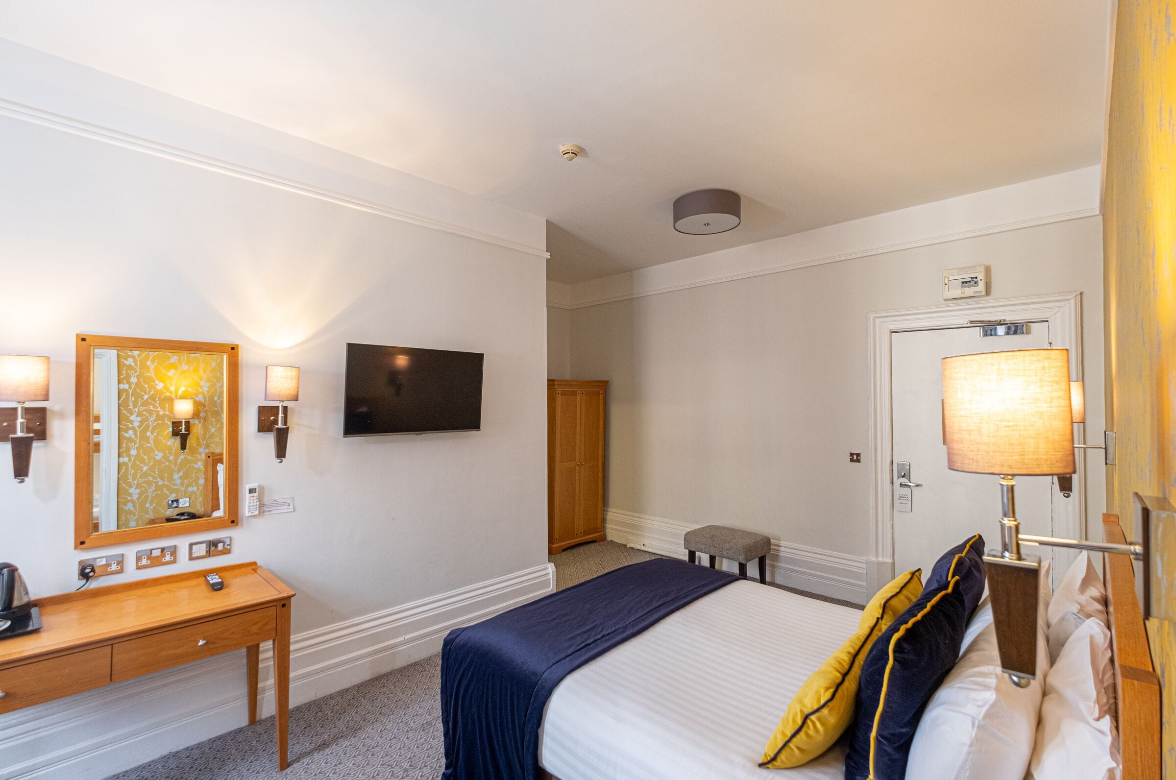 A hotel room with a bed, bedside table, lamp, TV, wardrobe, and a small stool.