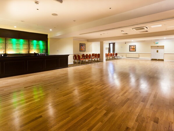 A spacious room with a wooden floor, a reception desk with a green-lit bar area, and rows of chairs. - The Durley Dean Hotel (Bournemouth)