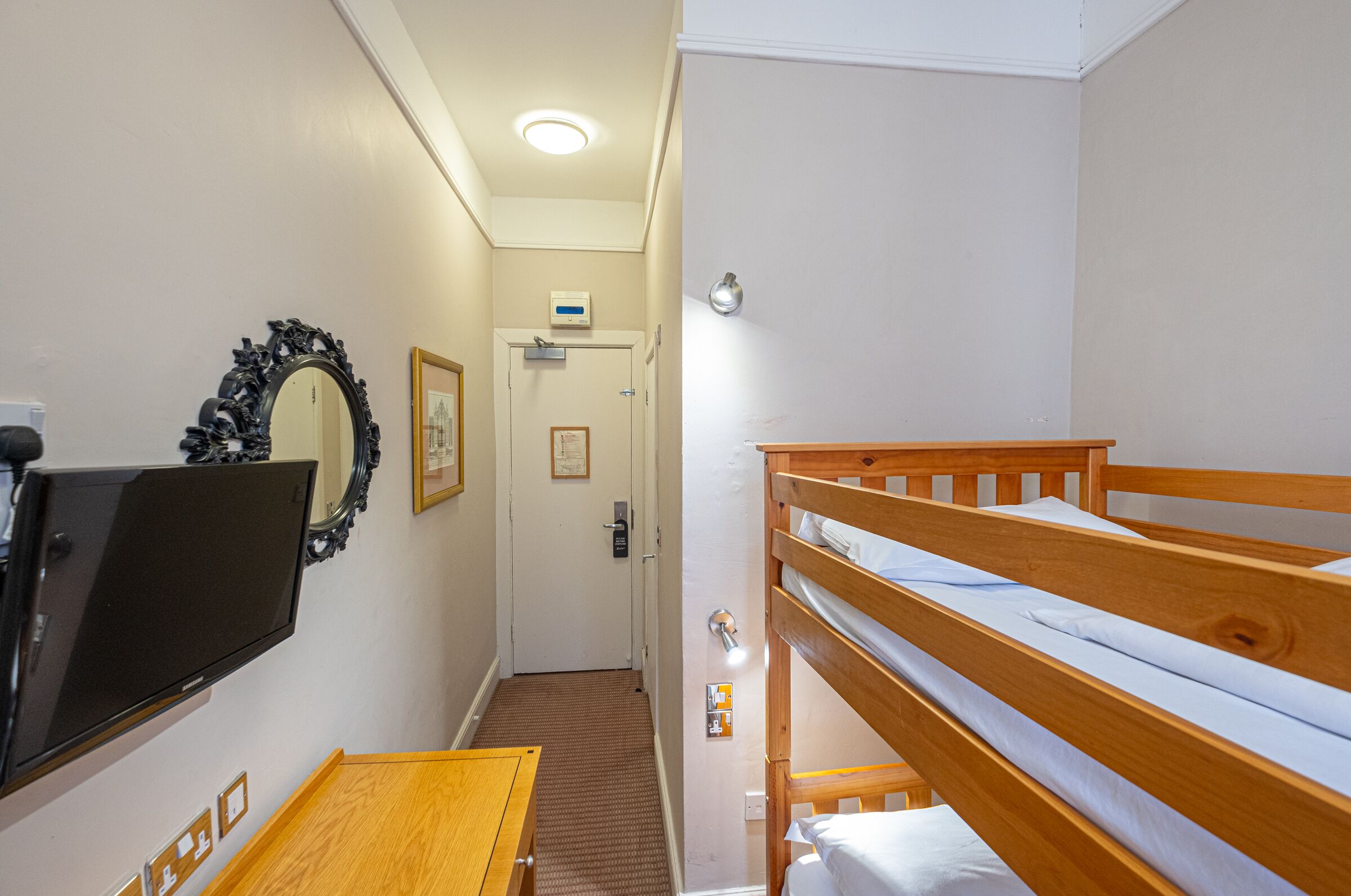Une chambre avec un lit superposé, un téléviseur à écran plat fixé au mur, une table en bois et une porte avec un interrupteur et une petite pancarte au-dessus.